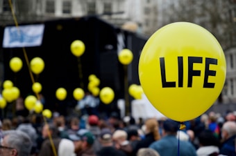A large group of people gathered outdoors, with several yellow balloons floating above the crowd. The most prominent balloon in the foreground has the word 'LIFE' printed in bold black letters.