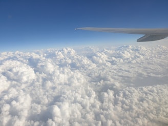 An airplane wing is visible against a backdrop of fluffy white clouds stretching across the sky. The vast expanse of clouds appears soft and dense, with a clear blue sky above.