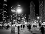 A bustling city street scene at night features blurred pedestrians in motion, surrounded by tall, brightly lit buildings. Traffic and street signs are visible, along with a prominent sign in the foreground encouraging membership in a nearby establishment.