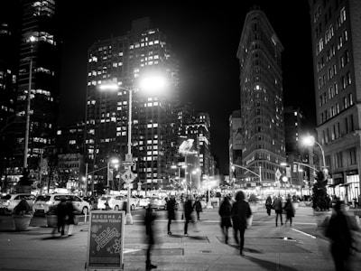 A bustling city street scene at night features blurred pedestrians in motion, surrounded by tall, brightly lit buildings. Traffic and street signs are visible, along with a prominent sign in the foreground encouraging membership in a nearby establishment.