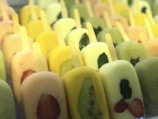 A variety of colorful popsicles are arranged in rows. Each popsicle contains pieces of fruit such as strawberries, kiwi, and possibly mint leaves. The colors range from green, yellow, to pale pink, suggesting different fruit flavors.