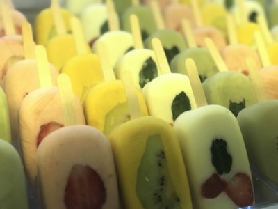 A variety of colorful popsicles are arranged in rows. Each popsicle contains pieces of fruit such as strawberries, kiwi, and possibly mint leaves. The colors range from green, yellow, to pale pink, suggesting different fruit flavors.