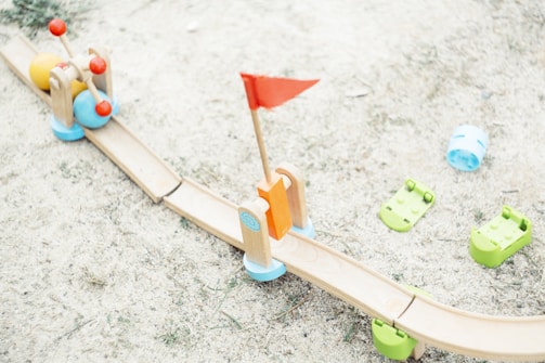 A wooden toy track on a sandy surface includes colorful toy balls and a red flag. The track is light brown with green bases, supplemented by playful elements like blue and green plastic components scattered around.