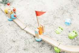 A wooden toy track on a sandy surface includes colorful toy balls and a red flag. The track is light brown with green bases, supplemented by playful elements like blue and green plastic components scattered around.