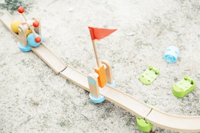 A wooden toy track on a sandy surface includes colorful toy balls and a red flag. The track is light brown with green bases, supplemented by playful elements like blue and green plastic components scattered around.