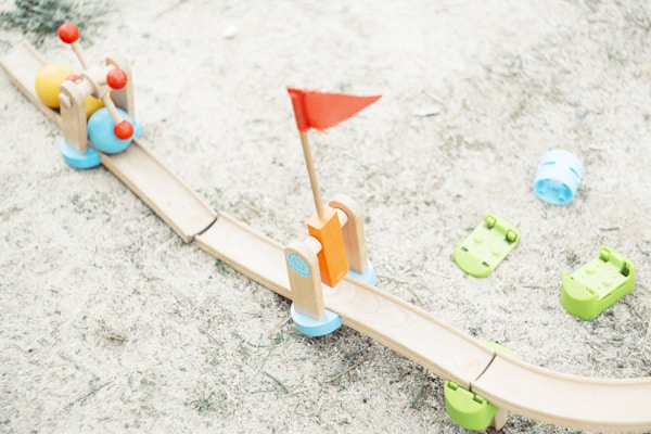 A wooden toy track on a sandy surface includes colorful toy balls and a red flag. The track is light brown with green bases, supplemented by playful elements like blue and green plastic components scattered around.