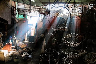 A sunlit workshop or market space filled with various metal objects and fixtures. Sunbeams filter through the overhead structure, casting shadows and creating a dramatic interplay of light and dark. Two people sit towards the left, engaged in work amidst bags and materials.