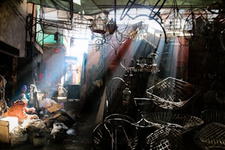 A sunlit workshop or market space filled with various metal objects and fixtures. Sunbeams filter through the overhead structure, casting shadows and creating a dramatic interplay of light and dark. Two people sit towards the left, engaged in work amidst bags and materials.