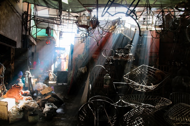 A sunlit workshop or market space filled with various metal objects and fixtures. Sunbeams filter through the overhead structure, casting shadows and creating a dramatic interplay of light and dark. Two people sit towards the left, engaged in work amidst bags and materials.