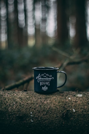 A black metal mug is placed on a horizontal tree trunk in a forest setting. The mug features the phrase 'The Adventure Begins' in white cursive lettering, with a mountain graphic above it.