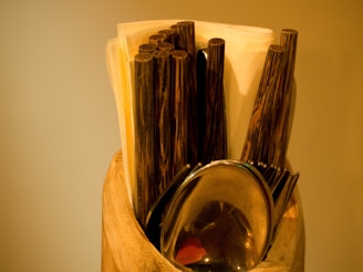 A collection of dark wooden chopsticks, a metal spoon, and folded napkins arranged in a holder with a warm, yellowish background.