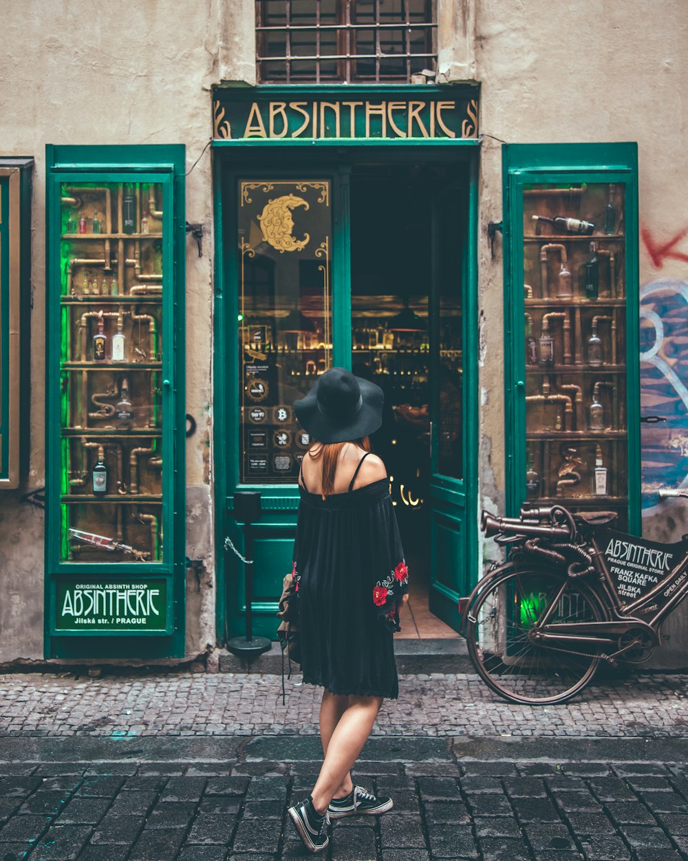woman in black off shoulder dress and black sun hat looking at green colored door building
