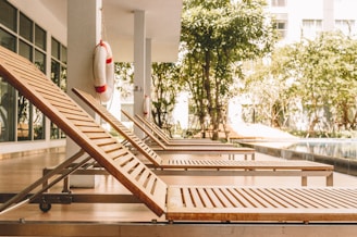 Several wooden lounge chairs are lined up along a poolside patio area, shaded by a building overhang. Life preservers are attached to the columns. There are trees and greenery in the background, adding a natural touch to the modern architecture.
