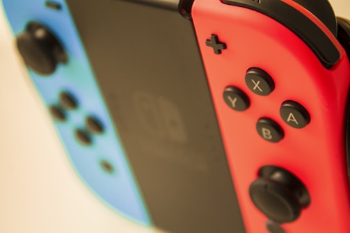 Close-up image of a gaming console with two detachable controllers. The left controller is blue with a black joystick and buttons, while the right controller is red with the same features. The central screen is slightly blurred in the background.