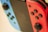 Close-up image of a gaming console with two detachable controllers. The left controller is blue with a black joystick and buttons, while the right controller is red with the same features. The central screen is slightly blurred in the background.