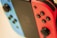 Close-up image of a gaming console with two detachable controllers. The left controller is blue with a black joystick and buttons, while the right controller is red with the same features. The central screen is slightly blurred in the background.