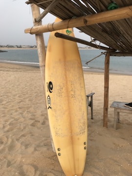 A surfboard is leaning against a wooden structure on a sandy beach, with a calm body of water in the background. The beach appears quiet and serene, with a distant view of a coastline. The wooden beams provide partial shade over the area, and a pair of sandals rests on a nearby bench.