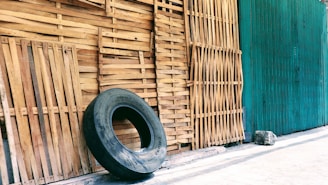 A large, worn-out tire leans against a wall made of vertically aligned wooden slats. The background is a mix of natural wood and a green-painted surface, with a concrete floor.