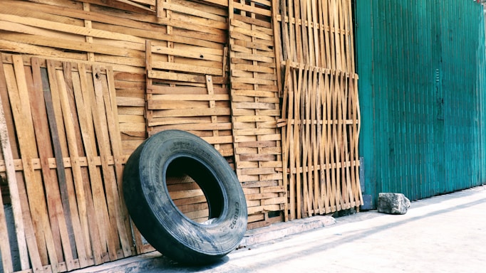 A large, worn-out tire leans against a wall made of vertically aligned wooden slats. The background is a mix of natural wood and a green-painted surface, with a concrete floor.
