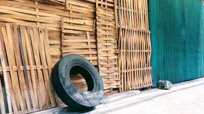 A large, worn-out tire leans against a wall made of vertically aligned wooden slats. The background is a mix of natural wood and a green-painted surface, with a concrete floor.