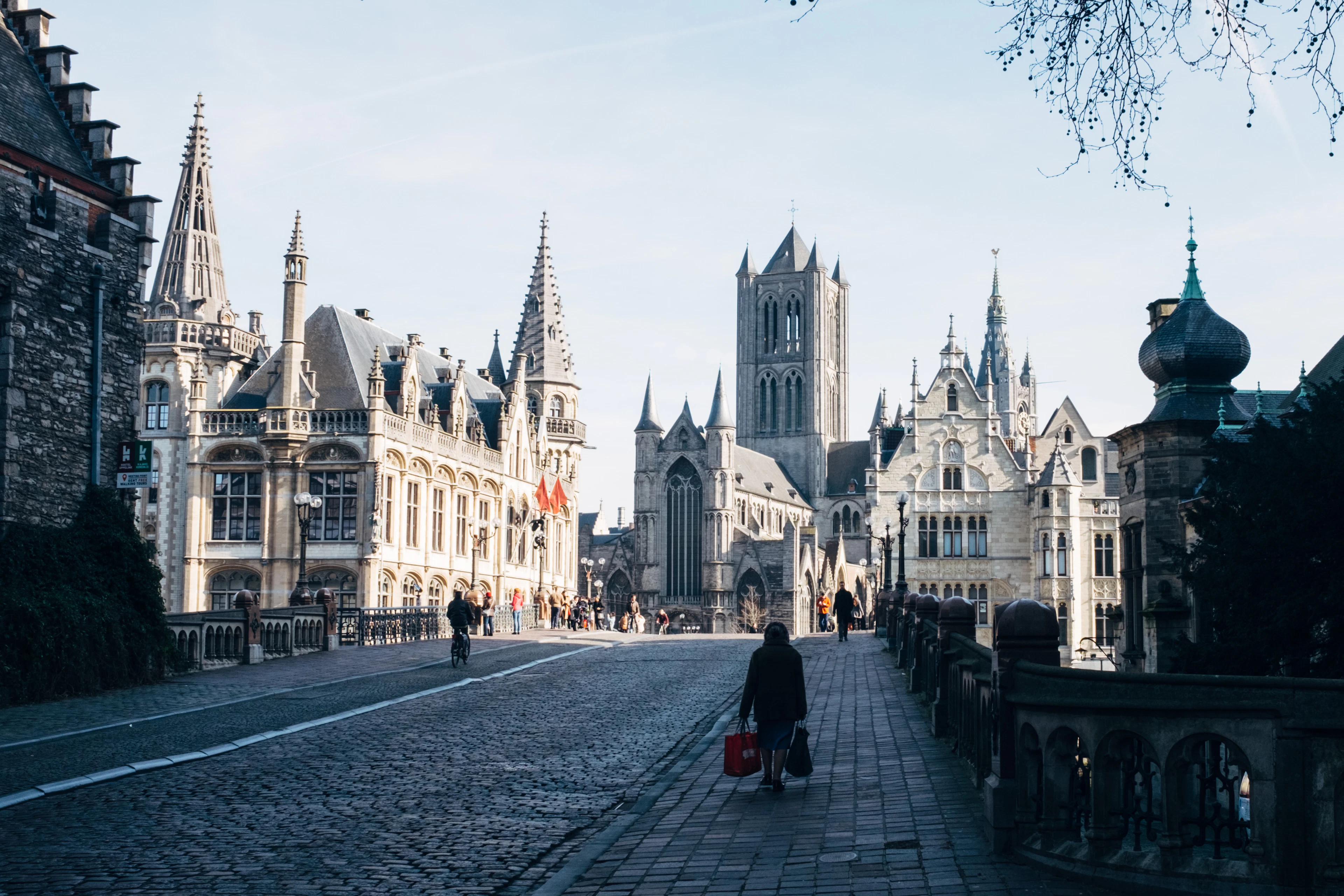 The cobbled Saint Michael's Bridge in Ghent city centre looking towards Saint Nicholas' Church and the Belfry, a lone figure with a red bag walking in low winter sun
