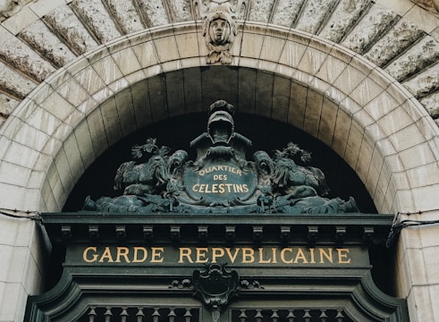 An ornate stone archway features intricate carvings and sculptural elements, with the words 'Garde Républicaine' prominently displayed in gold against a dark backdrop. Above this, the phrase 'Quartier des Celestins' is inscribed within a decorative shield, flanked by sculpted figures of horses, conveying a sense of history and grandeur.