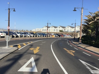 A wide road curves to the right with clearly marked lanes and directional arrows. A parking area filled with numerous cars lines the left side of the road. Lamp posts flanking the road have a vintage design. In the background, multiple neatly arranged white and grey buildings are visible, resembling residential or commercial structures. Palm trees and other greenery are present along the right side of the road.