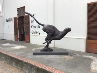 A sculpture of a cheetah captured in mid-run is displayed on a rectangular pedestal. The artwork is situated in front of an art gallery with white walls and wooden doors. The ground appears to be made of concrete and brick, and there is text on the wall that reads 'ART on Church Gallery' and 'Nature'.