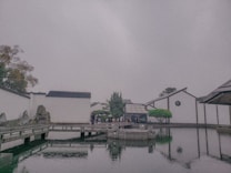 The image depicts a serene landscape featuring traditional architectural elements and a reflecting pool. White walls with black tile roofs are seen, typical of classical Chinese aesthetics. A wooden bridge crosses over the water, leading to a central structure surrounded by neatly arranged rocks and pruned greenery.