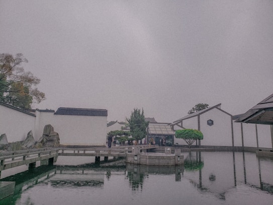 The image depicts a serene landscape featuring traditional architectural elements and a reflecting pool. White walls with black tile roofs are seen, typical of classical Chinese aesthetics. A wooden bridge crosses over the water, leading to a central structure surrounded by neatly arranged rocks and pruned greenery.