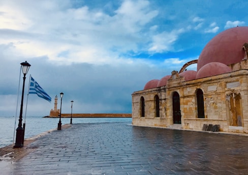 An old stone building with several domes stands by a waterfront. A Greek flag waves beside a lamppost on a wet cobblestone path, leading to a lighthouse in the distance. The sky is cloudy with patches of blue.