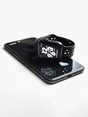 A sleek, black smartwatch with a sporty strap is resting on top of a black smartphone. The watch face displays the time with bold white numbers and additional information in smaller text. Both devices have a glossy finish that reflects light.
