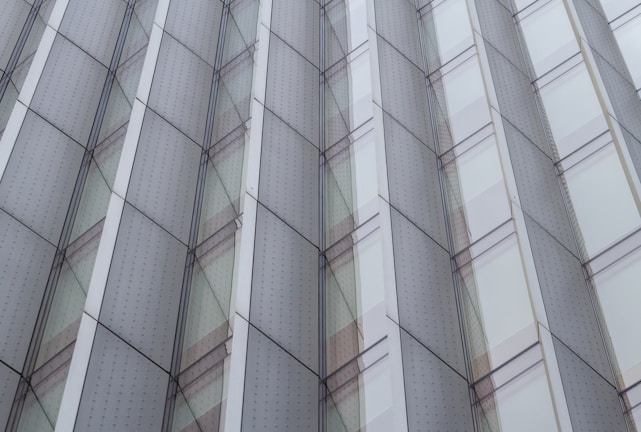 A modern architectural facade featuring repeating geometric patterns with glass panels and metallic frames. The design exhibits a symmetrical and orderly arrangement, creating a sense of depth and reflection.