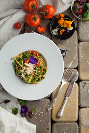 A plate of pasta garnished with a purple edible flower is surrounded by fresh ingredients, including whole tomatoes on the vine and assorted colorful edible flowers. There is a dish of flowers and leafy greens on the side, along with a fork and knife set on a stone slab surface.