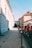 A group of cyclists wearing red outfits ride down a street alongside historical buildings with classical architectural features. The sky is clear, and the road is flanked by evenly spaced black bollards. Sunlight casts long shadows, adding to the visual depth of the scene.