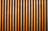 Vertical wooden slats evenly spaced with a dark background between them, creating a pattern of alternating light and dark stripes. The wood appears smooth and polished, with visible natural grain and knots.