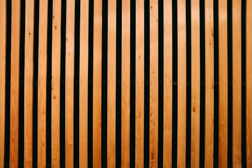 Vertical wooden slats evenly spaced with a dark background between them, creating a pattern of alternating light and dark stripes. The wood appears smooth and polished, with visible natural grain and knots.
