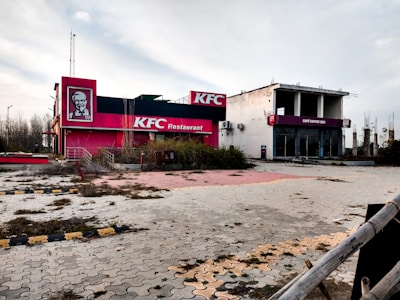 An abandoned fast-food restaurant and cafe, with overgrown vegetation and signs of neglect. The building features faded branding with a red and white color scheme, indicative of a well-known chicken franchise. The area appears desolate with cracked pavement and remnants of vegetation.