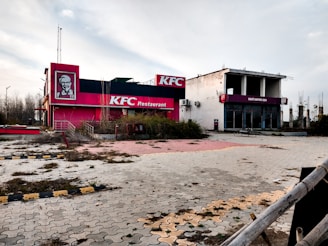 An abandoned fast-food restaurant and cafe, with overgrown vegetation and signs of neglect. The building features faded branding with a red and white color scheme, indicative of a well-known chicken franchise. The area appears desolate with cracked pavement and remnants of vegetation.