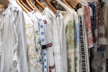 A row of intricately embroidered blouses hanging on wooden hangers. Each garment features unique patterns in various colors such as blue, red, and green, set against a predominantly white fabric. The embroidery includes floral and geometric designs.