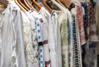 A row of intricately embroidered blouses hanging on wooden hangers. Each garment features unique patterns in various colors such as blue, red, and green, set against a predominantly white fabric. The embroidery includes floral and geometric designs.
