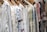 A row of intricately embroidered blouses hanging on wooden hangers. Each garment features unique patterns in various colors such as blue, red, and green, set against a predominantly white fabric. The embroidery includes floral and geometric designs.