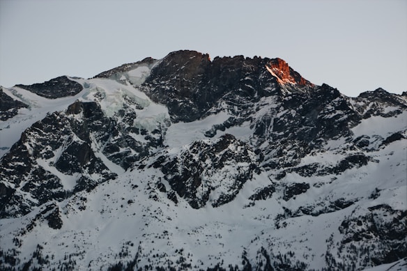 A rugged, snow-covered mountain peak is highlighted by the warm glow of sunrise or sunset. The rocky surfaces are interspersed with patches of snow, creating a stark contrast against the clear sky.