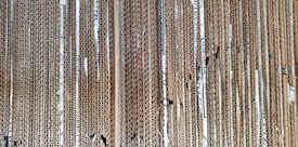 A stack of vertically aligned corrugated cardboard sheets with visible edges, showing the wavy texture within the layers. The cardboard appears densely packed, and some sheets show wear and tear with small gaps and bends.