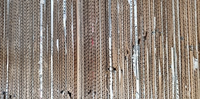 A stack of vertically aligned corrugated cardboard sheets with visible edges, showing the wavy texture within the layers. The cardboard appears densely packed, and some sheets show wear and tear with small gaps and bends.
