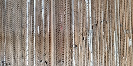 A stack of vertically aligned corrugated cardboard sheets with visible edges, showing the wavy texture within the layers. The cardboard appears densely packed, and some sheets show wear and tear with small gaps and bends.
