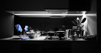 A kitchen counter is under a bright, focused light. Several kitchen items including pots, a metal bowl with utensils, wine glasses, and a cup are neatly arranged. A plant is placed near the right side, adding a touch of greenery. A bottle of wine and various kitchen tools are apparent, with a dish towel draped over some items. The overall setting suggests a modern kitchen with monochrome tones and a splash of blue from the towels.