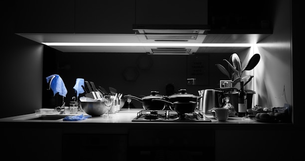 A kitchen counter is under a bright, focused light. Several kitchen items including pots, a metal bowl with utensils, wine glasses, and a cup are neatly arranged. A plant is placed near the right side, adding a touch of greenery. A bottle of wine and various kitchen tools are apparent, with a dish towel draped over some items. The overall setting suggests a modern kitchen with monochrome tones and a splash of blue from the towels.