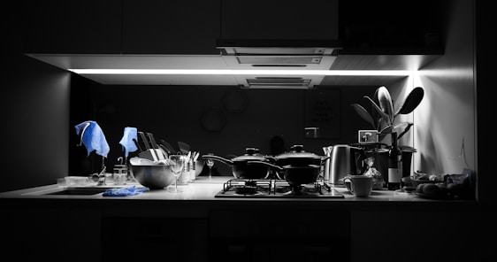 A kitchen counter is under a bright, focused light. Several kitchen items including pots, a metal bowl with utensils, wine glasses, and a cup are neatly arranged. A plant is placed near the right side, adding a touch of greenery. A bottle of wine and various kitchen tools are apparent, with a dish towel draped over some items. The overall setting suggests a modern kitchen with monochrome tones and a splash of blue from the towels.
