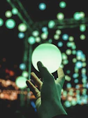 Close-up of a hand reaching toward a shimmering, translucent orb of light hovering in darkness.